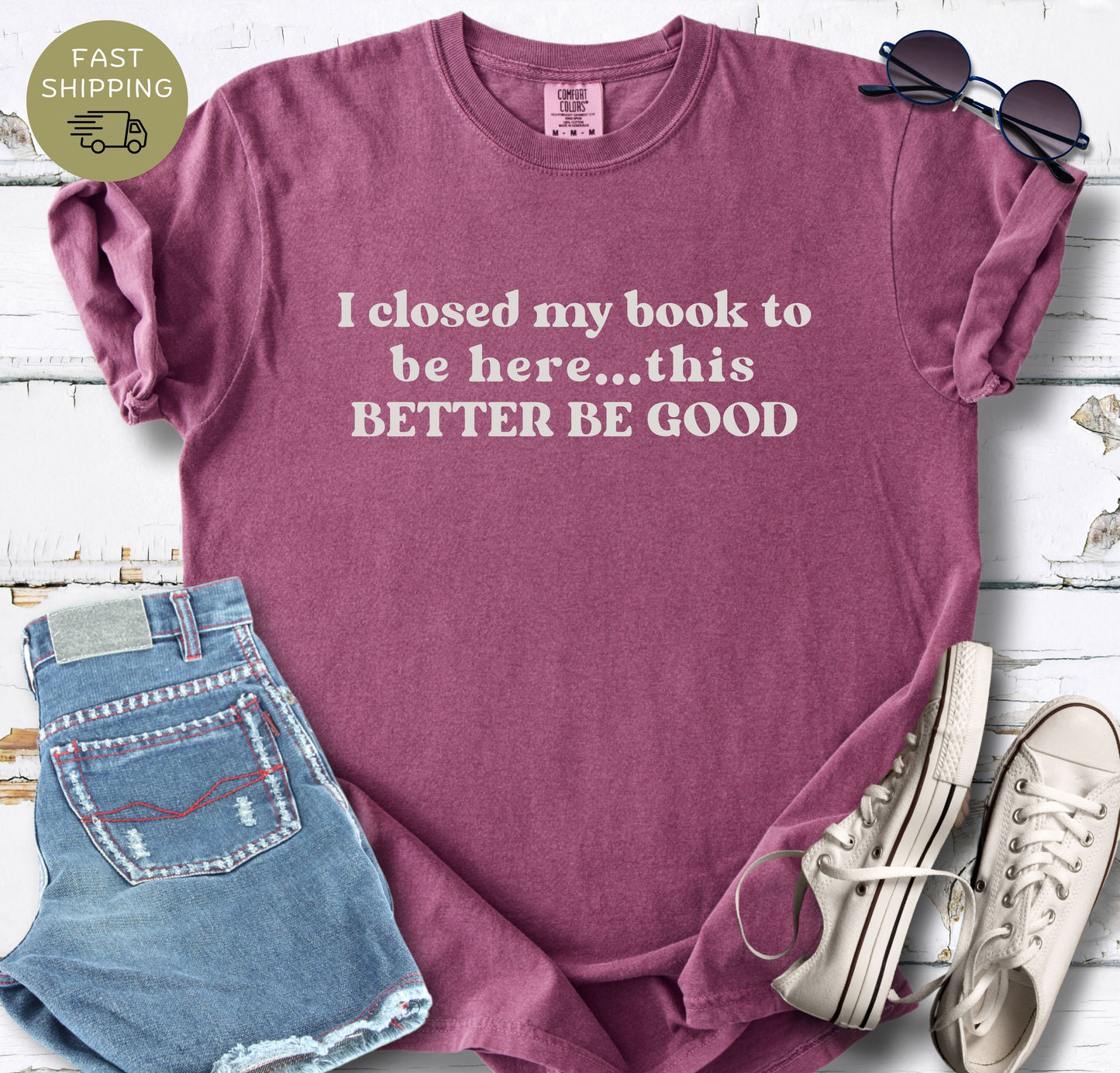 A flat-lay photo of a berry-colored t-shirt featuring the text "I closed my book to be here... this BETTER BE GOOD" in cream-colored vintage typography. The shirt is styled on a white wooden background next to denim shorts, white sneakers, and  sunglasses.
