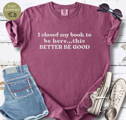 A flat-lay photo of a berry-colored t-shirt featuring the text "I closed my book to be here... this BETTER BE GOOD" in cream-colored vintage typography. The shirt is styled on a white wooden background next to denim shorts, white sneakers, and  sunglasses.