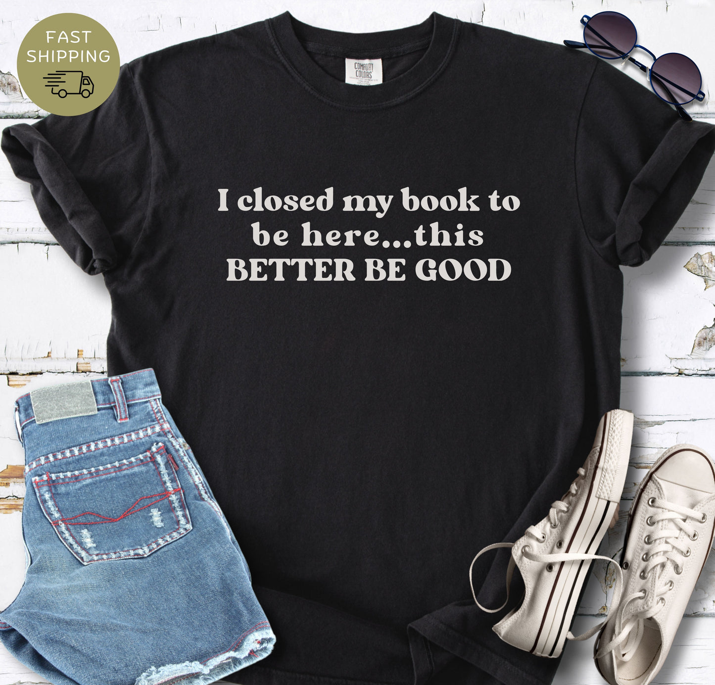 A flat-lay photograph of a Black graphic t-shirt featuring the text "I closed my book to be here... this BETTER BE GOOD" in a cream-colored vintage font, styled with denim shorts and white sneakers on a white wood background.