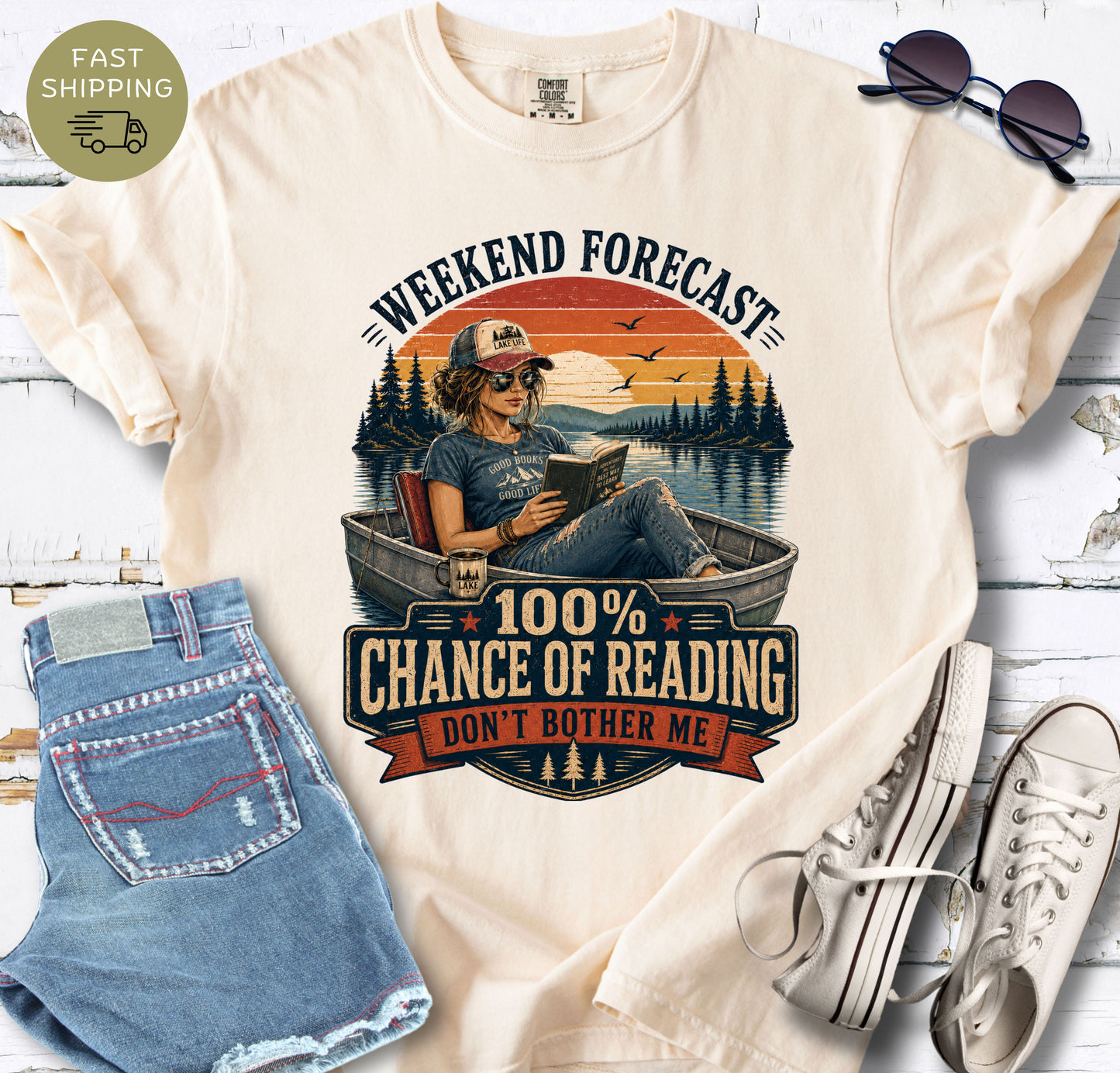 Book lover graphic tee showing a woman reading in a small boat on a calm lake at sunset.