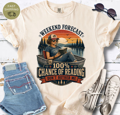 Book lover graphic tee showing a woman reading in a small boat on a calm lake at sunset.