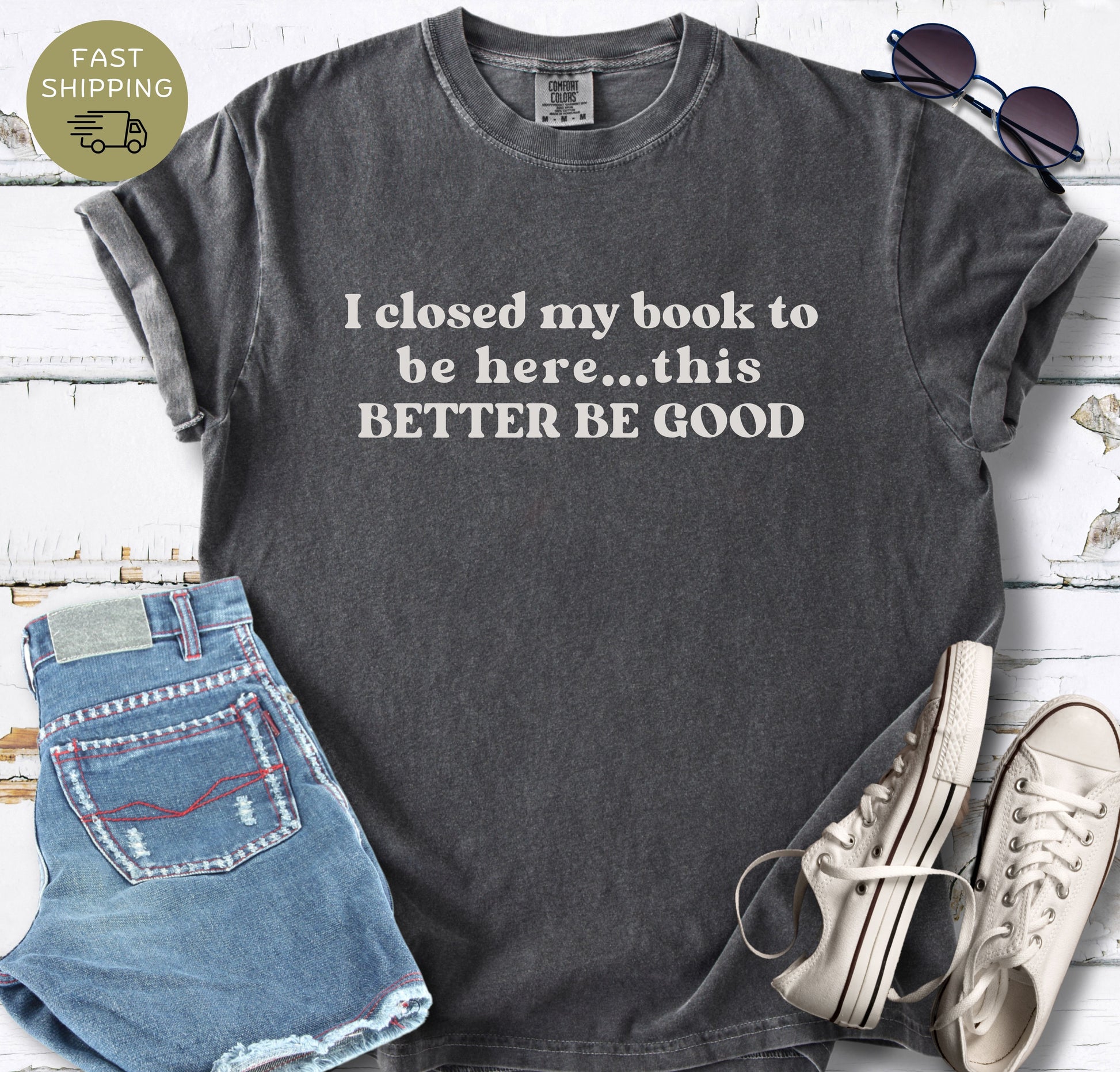 High-angle shot of a Pepper  t-shirt with "I closed my book to be here" printed on the chest. Styled with minimalist accessories on a white plank surface.